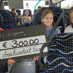 Kinder halten ein Schild mit 300.000 Euro auf einem Bus. Eines der Mädchen lächelt, andere sehen zu.