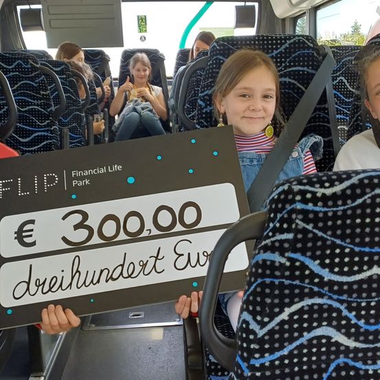 Kinder halten ein Schild mit 300.000 Euro auf einem Bus. Eines der Mädchen lächelt, andere sehen zu.