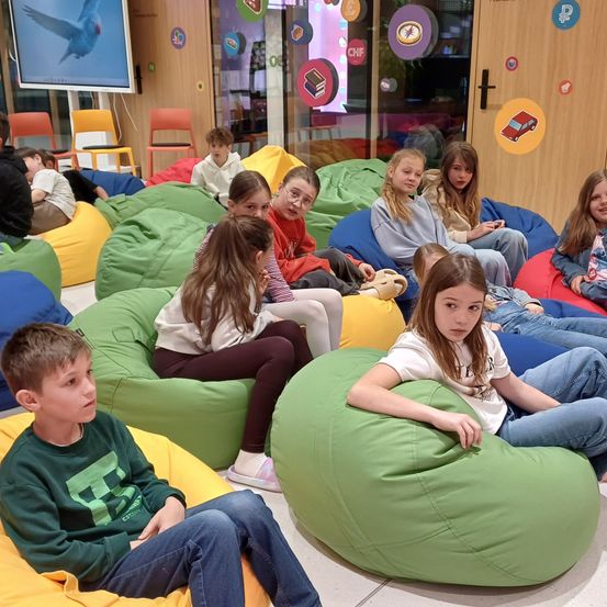 Eine Gruppe von Kindern sitzt auf Sitzsäcken in einem Raum mit einer Projektionsleinwand. Einige tragen eine Brille. Der Raum hat farbige Sitzsäcke und Holztüren mit Symbolen.