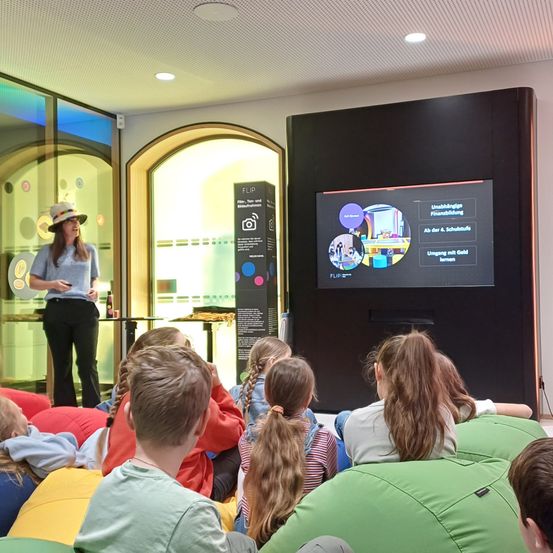 Eine Frau in einem Hut hält eine Präsentation für Kinder, die auf Beanbags vor einem Bildschirm sitzen. Der Bildschirm zeigt Inhalte zur Finanzerziehung.