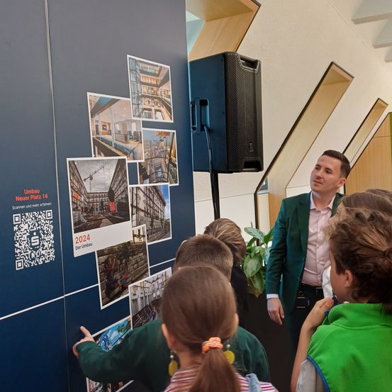 Ein Mann erklärt Kindern eine Präsentation über ein neues Stadtprojekt. Es gibt Bilder an der Wand und einen Lautsprecher. Die Kinder schauen sich die Präsentation an.