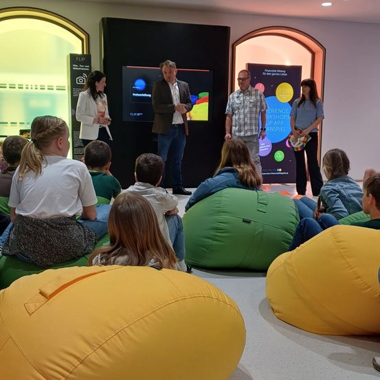Eine Gruppe von Menschen sitzt auf farbigen Sitzsäcken in einem Raum. Drei Personen stehen vor einem Bildschirm, möglicherweise bei einer Präsentation. Einer hält einen Becher und ein Stück Papier. Dahinter stehen zwei Frauen neben einem Banner.