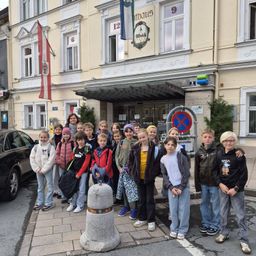 Eine Gruppe von Kindern steht auf einem Bürgersteig vor einem Gebäude mit Flagge und '12 Rathaus' Schild. Ein Auto ist in der Nähe geparkt.