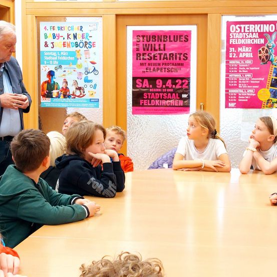 Ein Mann steht vor einem Klassenzimmer voller Kinder und bespricht etwas. Zwei Poster hängen an der Wand hinter ihnen.