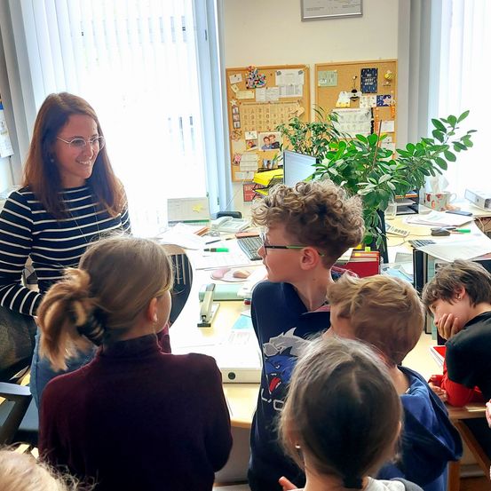 Eine Frau steht hinter einem Schreibtisch und lächelt eine Gruppe von Kindern an. Sie befinden sich in einem Klassenzimmer mit Pflanzen und Bulletin Boards im Hintergrund.
