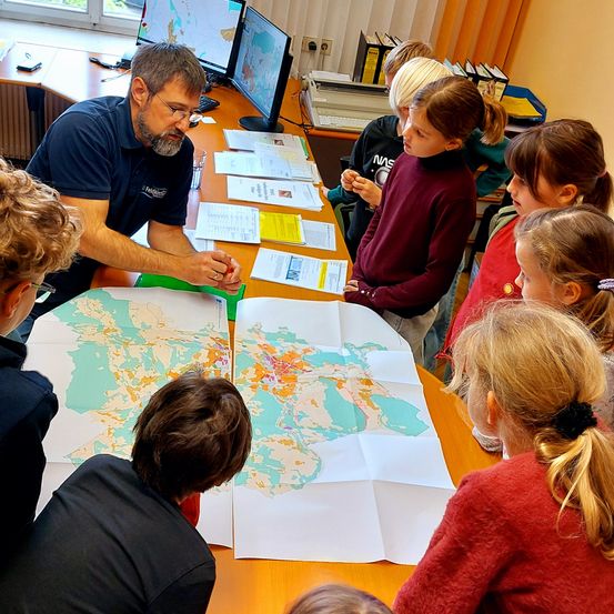 Eine Gruppe von Schülern sitzt um einen Tisch herum mit Karten und Papieren. Ein Mann mit Brille steht und erklärt etwas. Es gibt Computer und Bücher auf dem Schreibtisch.