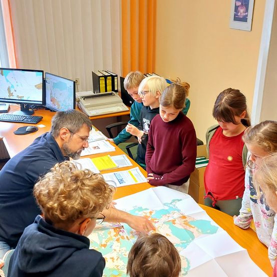 Eine Gruppe von Kindern und einem Mann versammelt sich um eine große Karte auf einem Tisch. Der Mann zeigt auf etwas auf der Karte, während die Kinder darauf schauen.