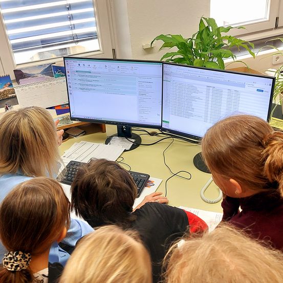 Eine Gruppe von Kindern sitzt in einem Klassenzimmer um einen Schreibtisch herum und schaut auf zwei Computerbildschirme. Eine Frau ist im Hintergrund.