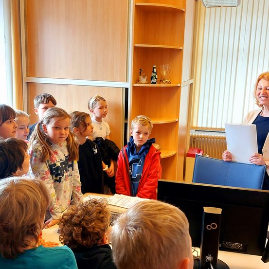 Eine Gruppe von Kindern steht um einen Schreibtisch und betrachtet ein von einer Frau gehaltenes Papier. Dahinter steht ein hölzerner Schrank, der mit verschiedenen Gegenständen gefüllt ist. Auf dem Schreibtisch befinden sich ein Monitor und ein Computer.