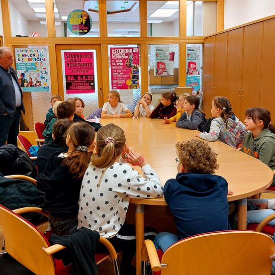 Eine Gruppe von Kindern sitzt um einen Tisch in einem Raum mit Plakaten an der Wand. Ein Erwachsener steht und spricht mit ihnen.