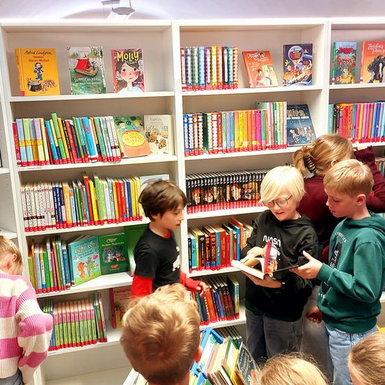 Mehrere Kinder stehen um ein Bücherregal herum, das mit bunten Büchern gefüllt ist. Ein Junge liest laut aus einem Buch vor, während andere aufmerksam zuhören. Die Bücher haben verschiedene Themen und bunte Cover.