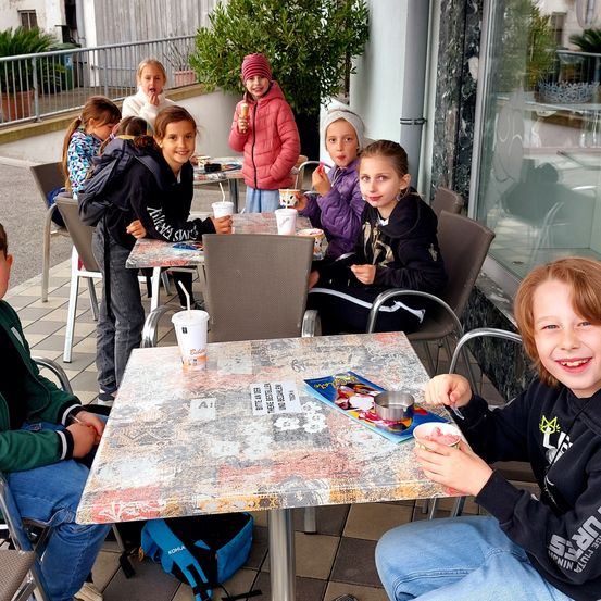 Eine Gruppe von Kindern sitzt draußen um einen Tisch herum und genießt Eis und andere Leckereien. Der Tisch hat ein farbenfrohes Design und die Kinder sind für das kalte Wetter gekleidet.