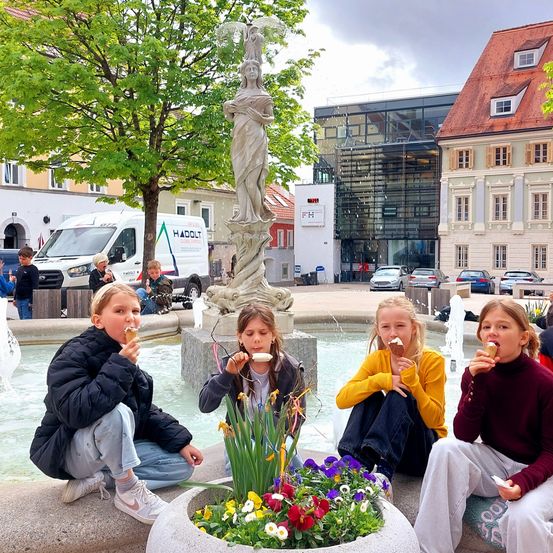 Vier Mädchen genießen Eis vor einem Brunnen auf einem Stadtplatz, umgeben von Gebäuden und einer Statue.