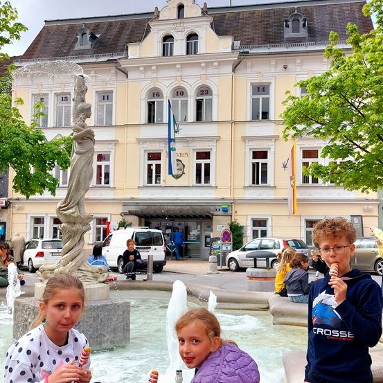 Drei Kinder in einem Brunnen, eines isst Eis, eine Statue, ein Van und ein Gebäude.