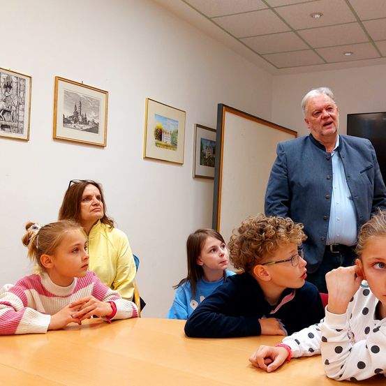 Eine Gruppe von Kindern und Erwachsenen sitzt um einen Tisch in einem Raum mit Bildern an der Wand. Ein älterer Mann steht hinter ihnen.