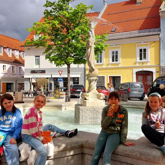 Vier junge Mädchen sitzen bei einem Brunnen auf einem Stadtplatz und essen Eis. Hinter ihnen befinden sich Gebäude mit roten Dächern und gelben Wänden. Ein Gebäude hat ein Schild mit der Aufschrift 'Il Custo'. Bäume und Autos sind sichtbar.
