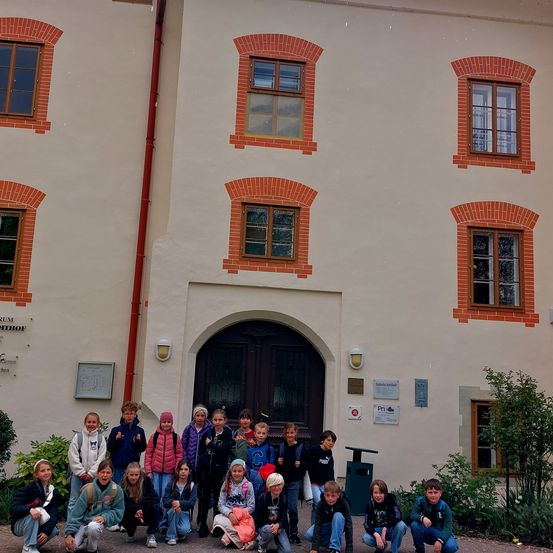 Eine Gruppe von Kindern posiert für ein Foto vor einem Gebäude mit mehreren Fenstern und einer Tür. Das Gebäude hat eine weiße Wand mit roten Ziegeldesigns.