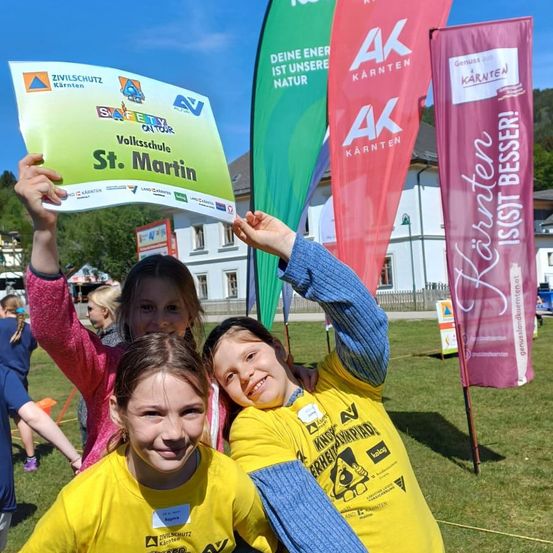 Drei Mädchen posieren für ein Foto und halten ein Schild für St. Martin, tragen gelbe T-Shirts. Dahinter sind Fahnen und ein weißes Gebäude unter einem blauen Himmel.