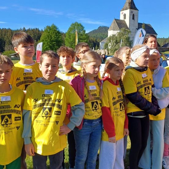 Eine Gruppe von Kindern in gelben T-Shirts steht draußen vor einer Kirche. Die gelben T-Shirts haben verschiedene Texte und Logos darauf. Eine Frau im Hintergrund schaut die Kinder an.