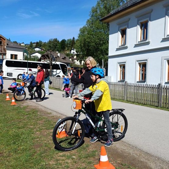 Eine Gruppe von Kindern fährt auf einem Pfad in der Nähe eines weißen Gebäudes mit Fahrrädern. Eine Frau führt ein Kind, das eine gelbe Jacke und einen blauen Helm trägt. Verkehrskegel markieren den Bereich.