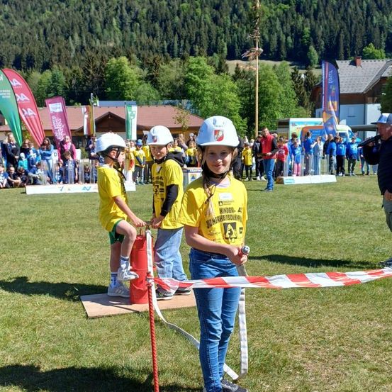 Drei Kinder in Helmen stehen auf einer Plattform und bereiten sich darauf vor, einen roten Seil zu ziehen. Sie sind von Zuschauern umgeben, und im Hintergrund sind Banner zu sehen.