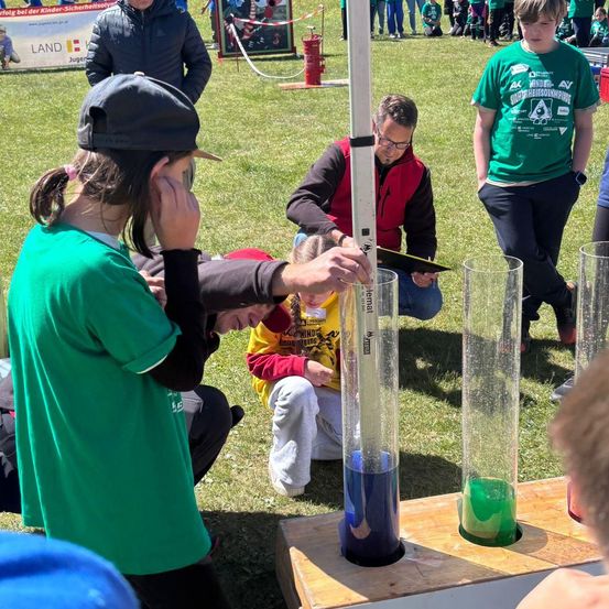 Schüler und Erwachsene beteiligen sich an einem Outdoor-Wissenschaftsexperiment. Ein Schüler in Grün hält einen Messstab, während ein anderer in Gelb kniet und beide beobachten farbige Flüssigkeiten in hohen Röhren.
