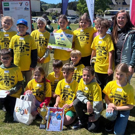 Eine Gruppe von Kindern in gelben T-Shirts posiert für ein Foto mit einer Trophäe und Zertifikaten. Eine Frau in einer Jacke steht neben ihnen.