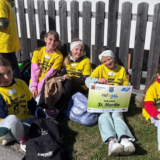 Eine Gruppe junger Mädchen in gelben Shirts sitzt auf Gras, hält ein grünes Schild mit St. Martin und lächelt.