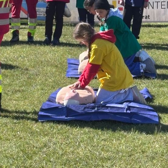 Eine Frau in einer gelb-roten Jacke übt CPR an einem Mannequin auf einem Rasen. Eine andere Frau beobachtet. Dahinter stehen Menschen in grünen und roten Uniformen.