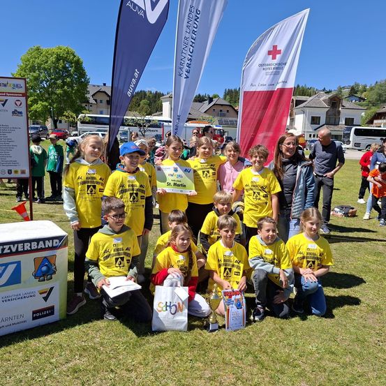 Eine Gruppe von Kindern in gelben T-Shirts posiert für ein Foto bei einer Veranstaltung im Freien. Eine Frau hält eine Urkunde, und ein Pokal liegt auf dem Rasen. Flaggen und ein Karton sind im Hintergrund zu sehen.