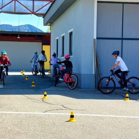 Eine Gruppe von Kindern fährt in einer Reihe auf Fahrrädern, mit Kegel auf dem Boden. Sie tragen Helme und Fahrradjacken. Ein Gebäude mit einer Garage und einer gelben Tür ist im Hintergrund.