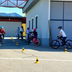 Eine Gruppe von Kindern fährt in einer Reihe auf Fahrrädern, mit Kegel auf dem Boden. Sie tragen Helme und Fahrradjacken. Ein Gebäude mit einer Garage und einer gelben Tür ist im Hintergrund.