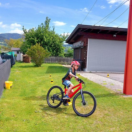 Ein Junge fährt auf einem Rasen mit Verkehrskegeln ein rotes Fahrrad. Dahinter befindet sich eine Garage mit einer Rolltor-Garagentür.