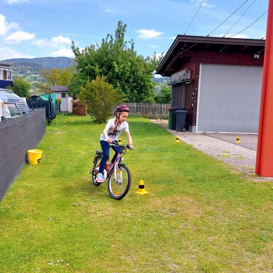 Ein Kind fährt auf einem Rasenstück mit einem gelben Eimer und Verkehrskegeln ein Fahrrad. Dahinter befindet sich eine Garage und ein Zaun, mit einer Bergkette in der Ferne.