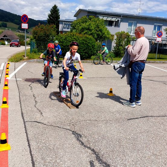 Eine Gruppe von Kindern fährt auf einem Parkplatz mit Kegel und einem Mann in der Nähe mit Fahrrädern. Die Kinder tragen Helme und Turnschuhe. Bäume und ein Haus sind hinter ihnen.