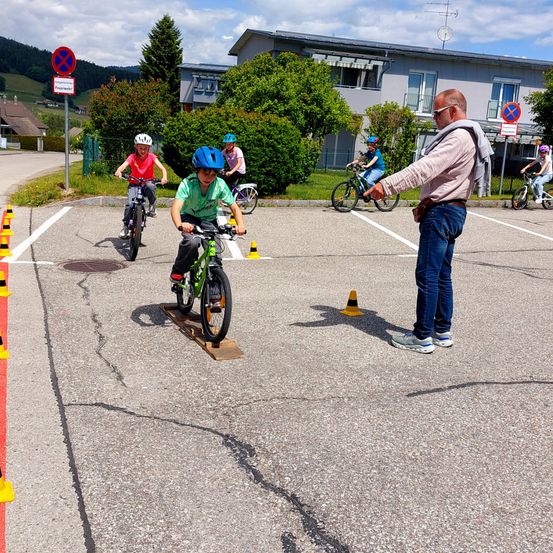 Ein Mann gibt Kindern auf Fahrrädern in einem Parkplatz mit Verkehrsschildern und Verkehrskegeln Anweisungen. Die Kinder üben das Fahren auf einer Rampe.