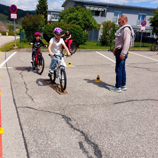 Drei Kinder fahren auf einem Parkplatz mit Fahrrädern. Ein Kind fährt auf einer Rampe. Ein erwachsener Mann steht in der Nähe, und zwei Fahrräder sind hinter einem Zaun geparkt.