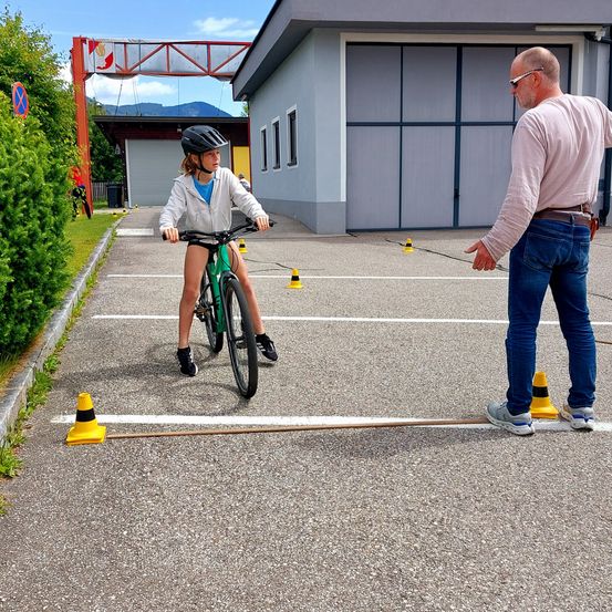 Ein junges Mädchen fährt ein Fahrrad auf einem Parkplatz, begleitet von einem Erwachsenen Mann. Um sie herum sind Kegel platziert.