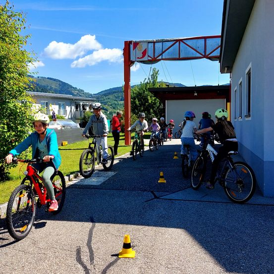 Mehrere Kinder mit Helmen fahren auf einem Betonweg mit Verkehrskegel auf Fahrrädern. Ein Gebäude mit Garage ist im Hintergrund zu sehen, zusammen mit einer Bergkette.