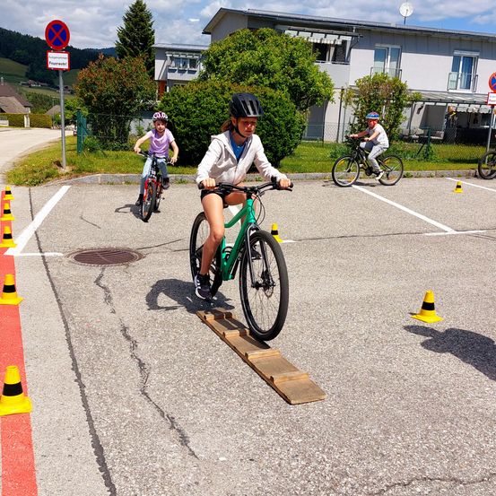 Drei Kinder fahren mit Fahrrädern auf einem Parkplatz. Das Mädchen vorne springt über eine Holzplanke. Sie tragen Helme.