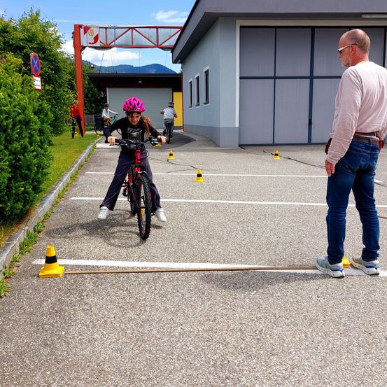 Ein junges Mädchen fährt zwischen gelben Kegel auf einer gepflasterten Fläche mit einem Mann. Hinter ihnen steht ein Gebäude mit großen Garagentoren.