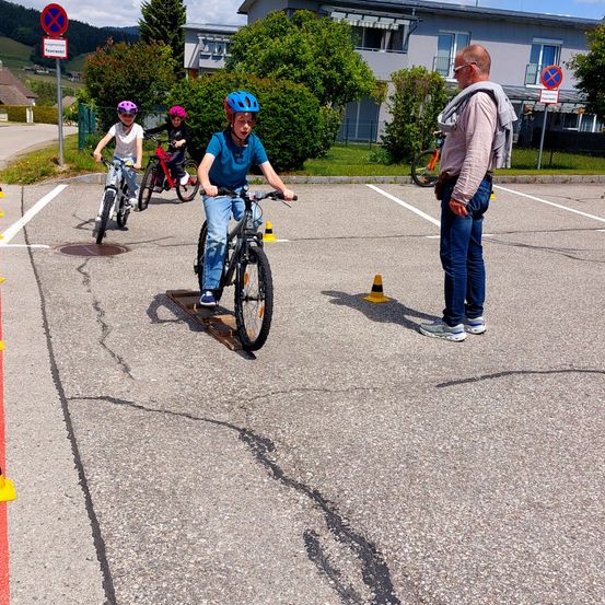 Drei Kinder mit Helmen fahren auf einem Parkplatz mit Fahrrädern. Ein Mann beobachtet sie von der Seite. Es gibt Verkehrsschilder, Büsche und ein Gebäude im Hintergrund.