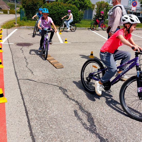 Kinder fahren auf einem Parkplatz mit Fahrrädern. Zwei von ihnen sind auf einer Holzplanke. Verkehrskegel sind aufgestellt. Ein weiteres Kind fährt hinter ihnen.