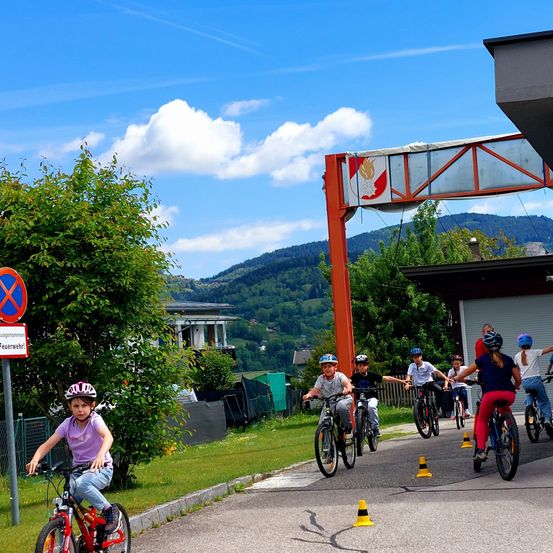 Eine Gruppe von Kindern fährt mit Fahrrädern auf einer Straße mit Verkehrskegeln und trägt Helme. Dahinter steht ein Gebäude mit einem roten Feuerzeichen. Im Hintergrund zeigt sich ein grasbewachsener Hügel mit einem Haus.