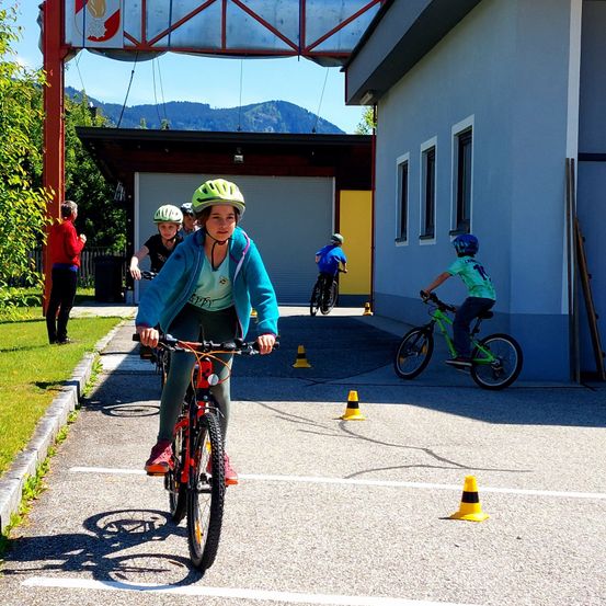Eine Gruppe von Kindern fährt mit Fahrrädern in einem Parkplatz mit Kegel. Ein Gebäude und Berge sind im Hintergrund.