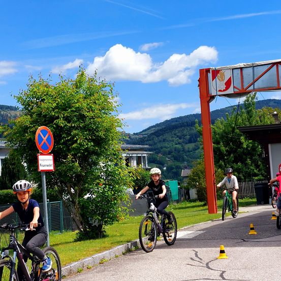 Eine Gruppe von Kindern fährt auf einer Straße mit Verkehrszeichen und einem Feuerwehrlogo mit Fahrrädern. Sie tragen Helme und scheinen für einen Wettbewerb zu üben.