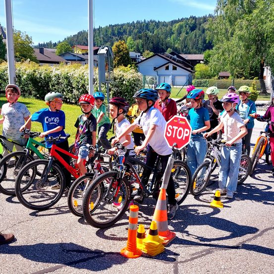 Eine Gruppe von Kindern steht mit ihren Fahrrädern in einer Reihe auf einem Parkplatz. Sie tragen alle Helme und es gibt Verkehrskegel und ein Stoppschild in der Nähe.