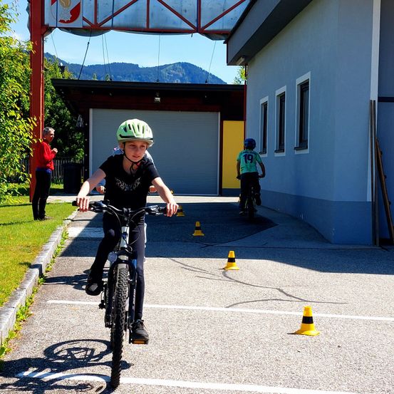 Ein junger Junge fährt mit einem Helm durch einen Parkplatz mit Hindernissen. Im Hintergrund fährt ein anderes Kind ein Fahrrad. Ein Gebäude mit gelber Wand ist sichtbar.