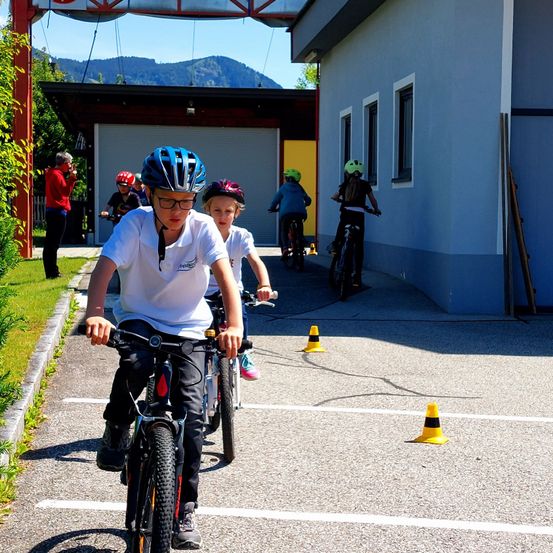 Kinder fahren mit Fahrrädern um Kegel auf einem gepflasterten Bereich vor einem Gebäude mit Garage. Berge sind im Hintergrund.