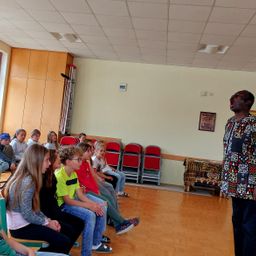 Ein Mann spricht laut zu einer Gruppe von Kindern, die auf Stühlen in einem Raum mit Holzboden und weißer Wand sitzen.